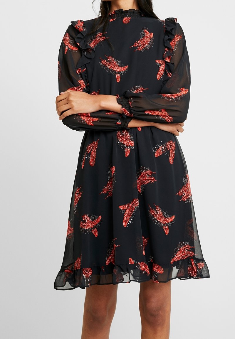 Robe noire à manches longues avec imprimé de plumes rouges, épaules et ourlet à volants, portée par une personne les bras croisés devant.