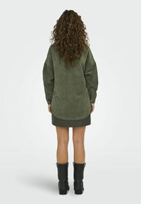Kobieta z długimi kręconymi włosami, ubrana w oversize'ową zieloną koszulę z korduroy, krótką zieloną spódniczkę i czarne botki do połowy łydki, stoi tyłem.