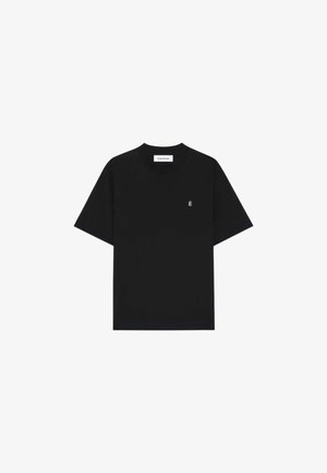 T-shirt noir à manches courtes et col rond avec un petit logo blanc "É" sur le côté gauche de la poitrine et une étiquette "Etudes Studio" à l'intérieur du col.