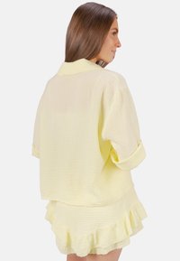 Chemise légère jaune en tissu texturé, avec des manches retroussées et une coupe décontractée, associée à une jupe volante.