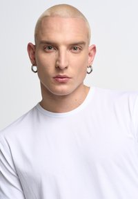 Witte crew-neck t-shirt, aansluitende silhouet, gedragen door een persoon met kort, lichtblond haar en ronde zilveren oorbellen.
