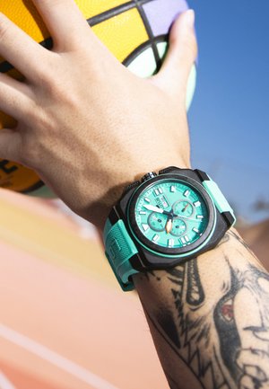 Roka z nošno turkizno športno uro s črnim ohišjem in tetovažo na zapestju drži pisano košarkarsko žogo na ozadju jasnega modrega neba.