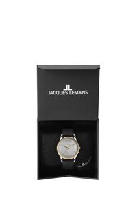 Sudraba pulkstenis ar zelta akcentiem, teksturēts pelēks ciparnieks, melns ādas josts, melnā prezentācijas kastē ar zīmola nosaukumu "JACQUES LEMANS."