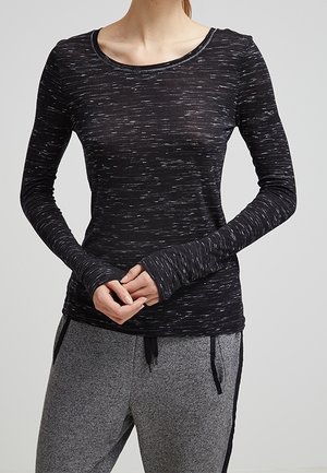 Vrouw met een zwart-wit gespikkeld top met lange mouwen met duimgaten en grijze joggingbroek met zwarte strepen aan de zijkant en trekkoorden.