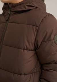 Rudas pūkinis švarkas su kapišonu, siūtu tekstūra ir užtrauktuku. Ant rankovės yra apvalus logotipo pleistras.