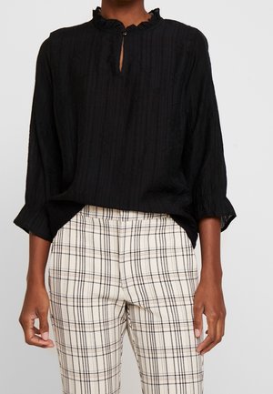 Osoba ubrana w czarną bluzkę z długim rękawem i falbaniastej kołnierzykiem oraz jasne spodnie w kratę, stojąca na tle jednolitego tła.