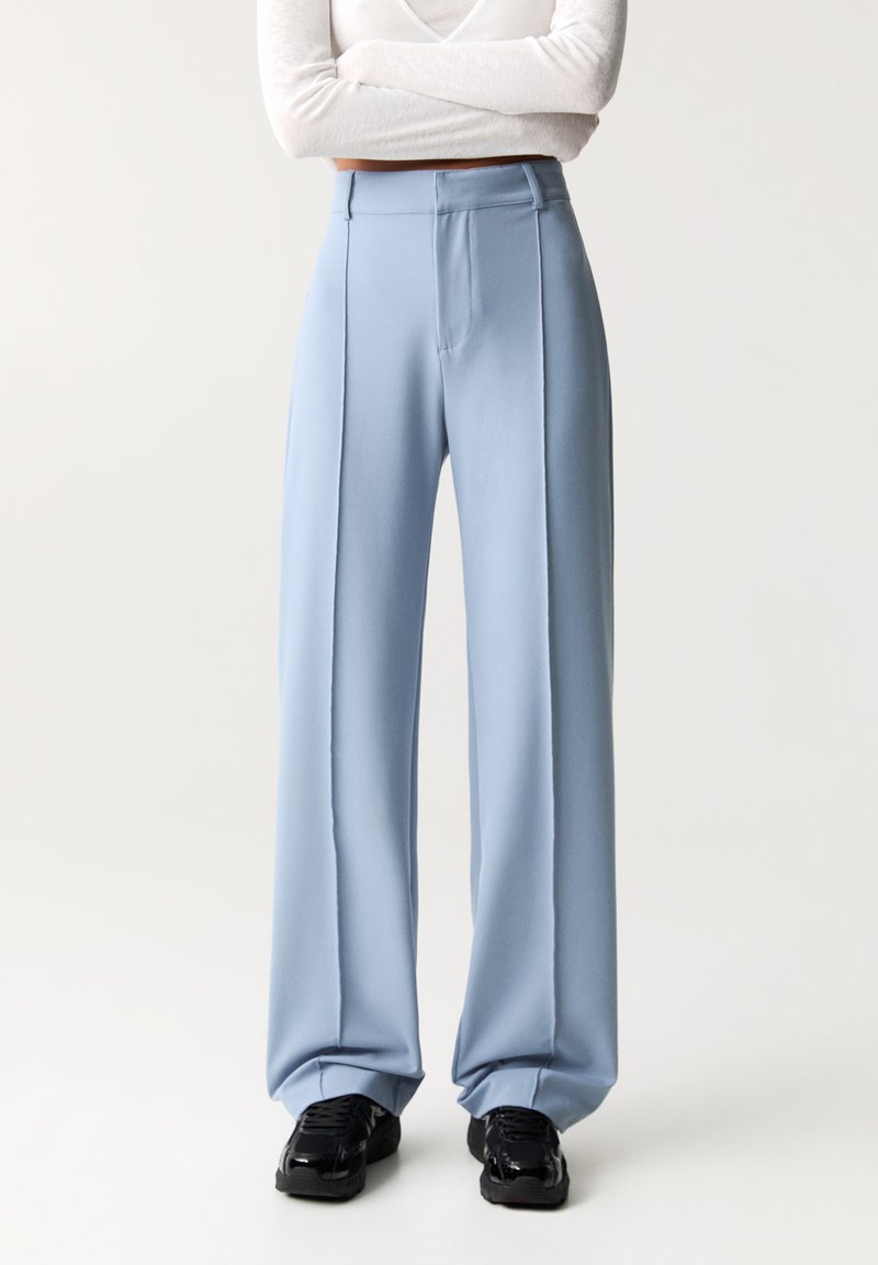 Hellblaue, weit geschnittene Hosen aus glattem Stoff, mit einer vorderen Nahtdetail und hohem Bund. Getragen mit schwarzen Sneakers.