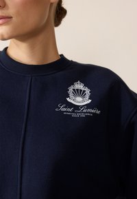 Felpa blu navy in morbido tessuto con logo ricamato bianco raffigurante una corona e il testo "Saint Lumière, Sporting Excellence, Since 1989."