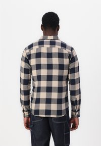 Man in een beige en marineblauwe geblokte lange mouwen shirt en donkere denim broek, staand met de rug naar de kijker tegen een witte achtergrond.