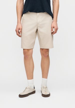 Man wearing beige knee-length shorts, white socks, cream sneakers, and a black shirt, standing against a plain light background.