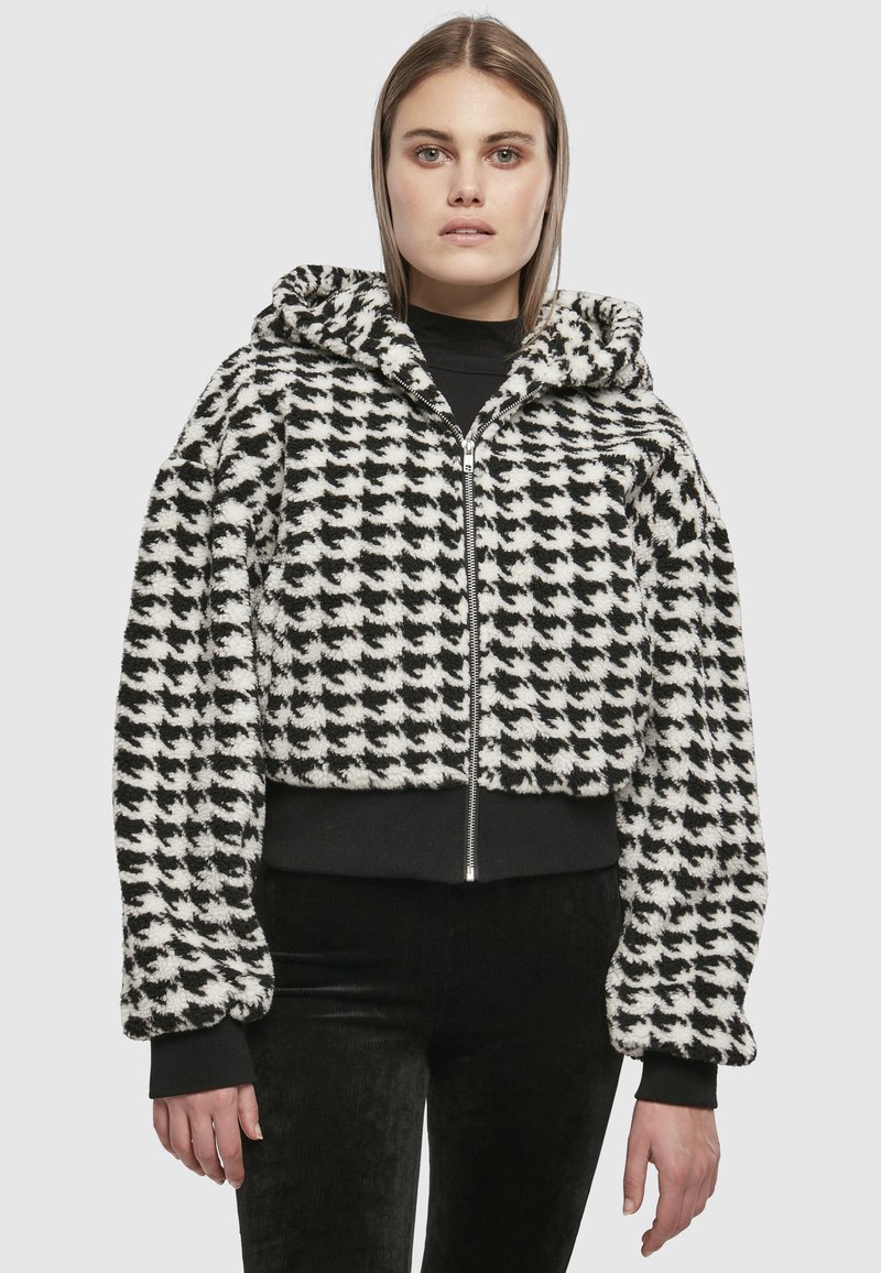 Femeie care poartă o jachetă din fleece cu model în carouri alb-negru, cu glugă, fermoar frontal și manșete negre, asortată cu pantaloni negri.