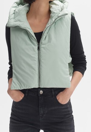 Personne portant une veste sans manches matelassée vert clair avec une fermeture éclair sur un t-shirt noir à manches longues et un jean noir, les mains dans les poches.