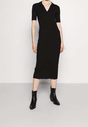 Woman wearing a black ribbed knit midi dress with short sleeves and a V-neck collar, paired with black ankle boots.