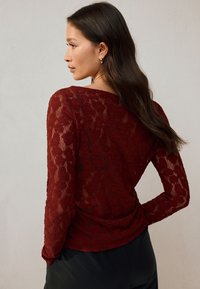 Top in pizzo color bordeaux con maniche lunghe, caratterizzato da un motivo floreale e tessuto a trama, abbinato a pantaloni neri. Vista posteriore mostrata.