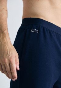 Pantalones cortos texturizados azul marino con una cinturilla ancha y un logo blanco bordado en el lado izquierdo. Mano colocada sobre la cinturilla.