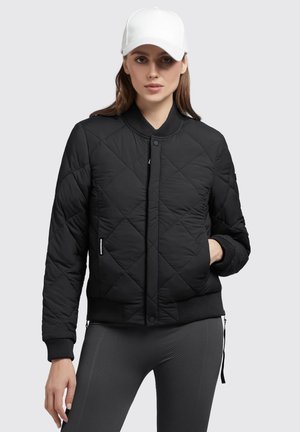Schwarze gesteppte Bomberjacke mit Rippstrickbündchen und Bund. Modell trägt eine weiße Mütze. Slim-Fit graue strukturierte Hose.