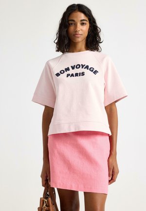 Kvinde iført lyserød "Bon Voyage Paris"-t-shirt og en mørkere lyserød nederdel, som holder en brun håndtaske mod en ensfarvet hvid baggrund.