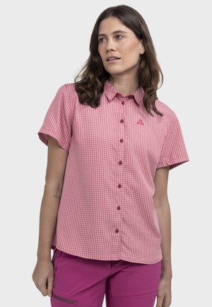 Frau trägt ein rosa kurzärmeliges Gingham-Karobluse mit Knöpfen und magentafarbene Hose, blickt leicht nach links.