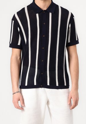 Homme portant un polo en maille à manches courtes bleu marine avec des rayures verticales blanches, une patte de boutonnage sur le devant, et un pantalon blanc, sur fond uni.