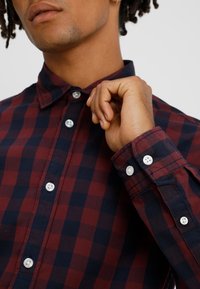 Lähikuva henkilöstä, joka on pukeutunut punavalkoraidalliseen kauluspaitaan, säätää kaulusta oikealla kädellään.