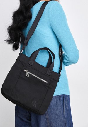 Kvinde iført blå sweater og denimbukser, bærende en sort Calvin Klein skuldertaske med lynlåslomme foran.