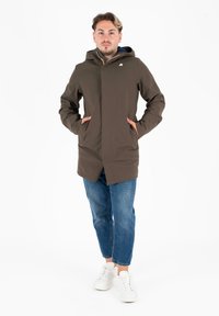Giacca impermeabile marrone con cappuccio, dotata di tasche laterali e logo. Abbinata a pantaloni in denim e sneakers bianche. Design semplice.