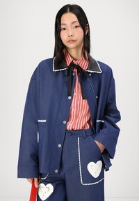 Denim jacket with a black collar and white trim, paired with a red and white striped shirt. Pants feature heart-shaped cutouts with white detailing.