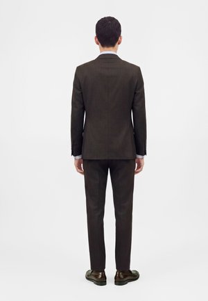Homme debout de dos portant un costume marron foncé avec un pantalon assorti et des chaussures marron sur un fond uni.