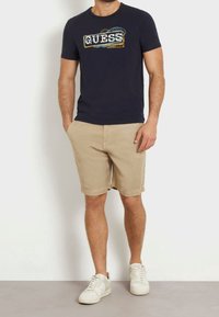 Camiseta azul marino con el logo de "GUESS", combinada con pantalones cortos beig y zapatillas blancas. Diseño sencillo y casual con una textura de tela suave.