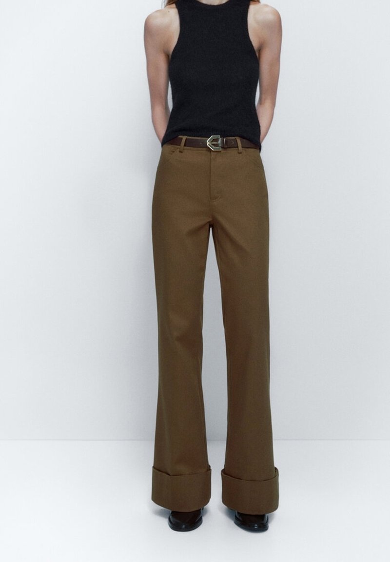 Brown wide-leg trousers with a high waist, a folded cuff detail, and a belt with a metallic buckle, paired with a black sleeveless top.