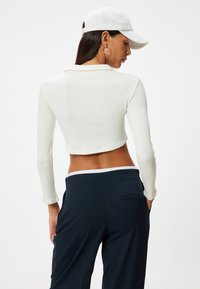 Weißes, geripptes, langärmliges, kurzes Oberteil und dunkle marineblaue Hose. Das Oberteil hat einen runden Halsausschnitt, und die Hose verfügt über Seitentaschen.