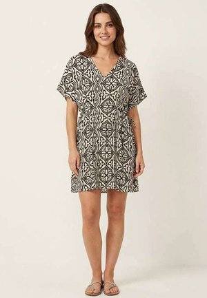 Femme debout et souriante, portant une robe courte à motifs noir et blanc et des sandales sur un fond uni.