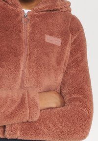 Sudadera rosa de felpa con cierre de cremallera y un tirador en forma de anillo. Cuenta con un pequeño parche de logo en el pecho y tejido texturizado.