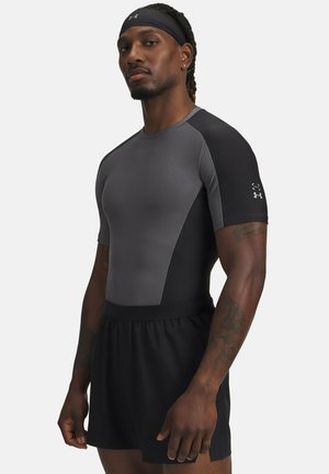 Athletischer Mann trägt ein grau-schwarzes, enganliegendes Sportshirt, schwarze Shorts und ein schwarzes Stirnband und steht mit neutralem Gesichtsausdruck.