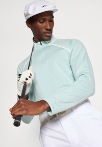 Gaiši zils garām piedurknēm golfa krekls ar puspogājamo apkakli, apvienots ar baltām biksēm un baltu plakanu cepuri. Uz rokas ir balts golfa cimdiņš.