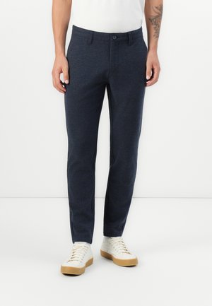 Pantalon bleu marine en tissu texturé avec une coupe slim, doté d'une fermeture à bouton et de poches latérales, associé à des tennis blanches.