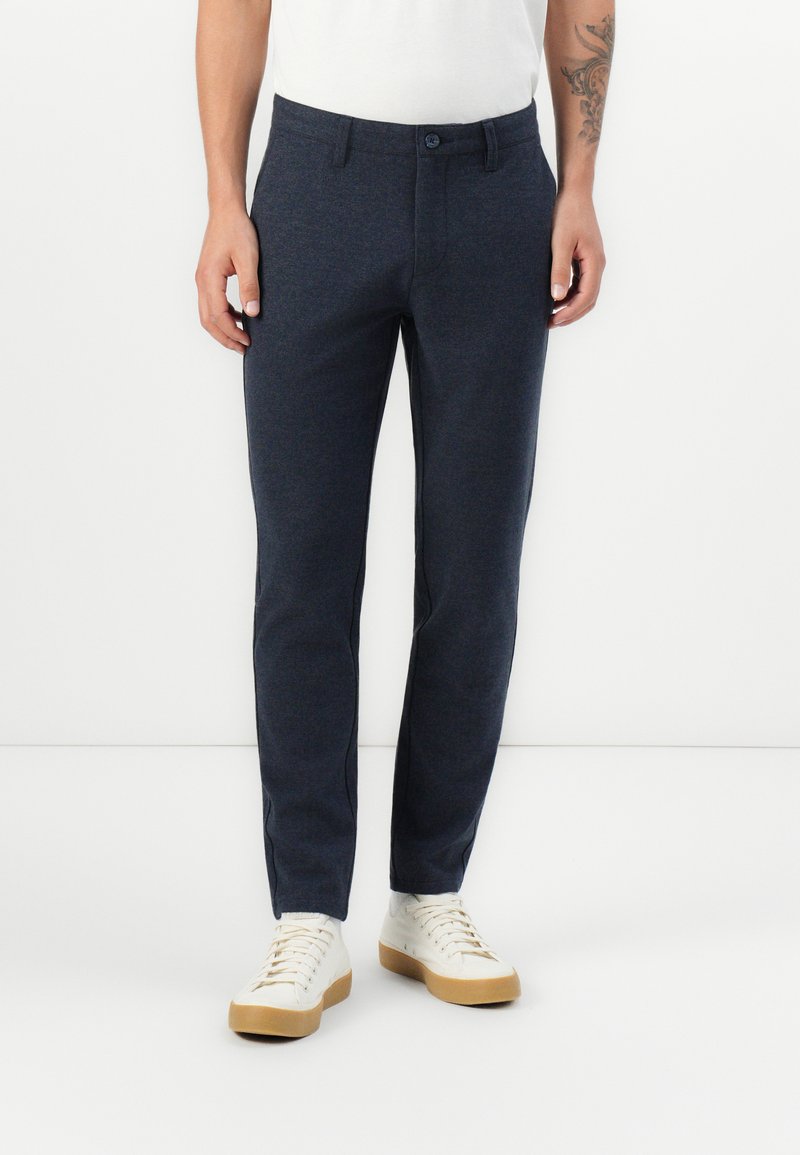 Pantalones azul marino de tejido texturizado con corte ajustado, que cuentan con un cierre de botón y bolsillos laterales, combinados con zapatillas blancas.