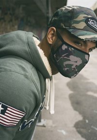 Masque de camouflage avec bord noir, porté avec un sweat à capuche vert doté d'une doublure en polaire blanche et de grandes lettres brodées. Casquette de camouflage.