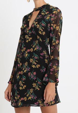 Vrouw die een zwarte bloemenjurk draagt met lange mouwen, een hoge hals met een sleutelgatuitstulping en gele, roze en groene bloemenpatronen.