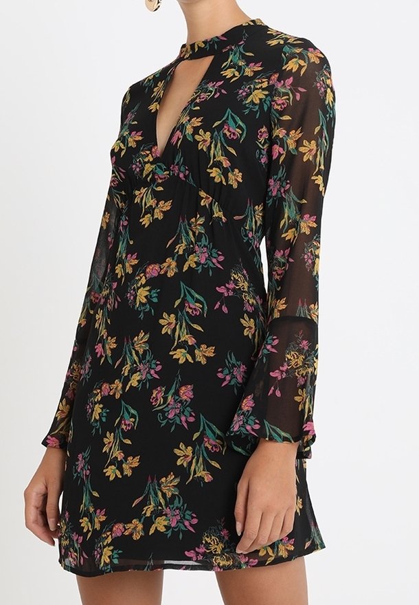 Femme portant une robe noire à motifs floraux avec manches longues, un col haut avec une découpe en forme de goutte, et des motifs floraux jaunes, roses et verts.