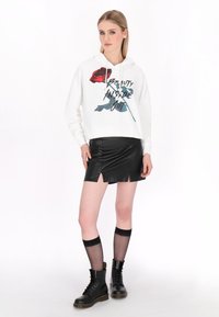 Bílá cropped mikina s grafickým prvkem červené růže a textem. Černá kožená mini sukně s předními rozparky. Modelka má na sobě černé šněrovací bojové boty.