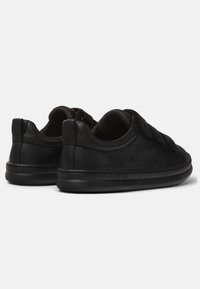 Des baskets en cuir noir présentent une texture lisse, une forme arrondie et des sangles élastiques. La semelle est noire avec un logo discret sur le côté.