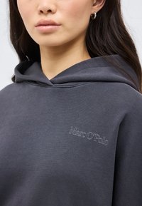 Νεαρό άτομο που φοράει σκούρα γκρι φούτερ με κουκούλα, με κεντημένο το λογότυπο "Marc O'Polo" στο στήθος, εμφανίζοντας τα χείλη, το πηγούνι και μέρος του αυτιού με ασημένιο σκουλαρίκι κρίκο.