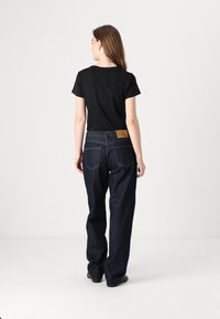 Camiseta negra de manga corta combinada con unos jeans de mezclilla oscura y holgados, que muestran un parche de cuero en la parte trasera de la cintura. Textura de tela suave.