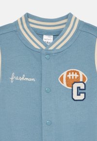 Helles blaues Varsity-Jacket mit cremefarbenem gestreiften Kragen, durchgängiger Knopfleiste, besticktem "Freshman", Fußball-Patch und dem Buchstaben "C" in Marineblau.