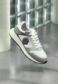 Chaussure de course légère comprenant une tige en mesh blanc et du daim gris, avec une semelle extérieure en caoutchouc noir texturé et un détail de logo sur le côté.