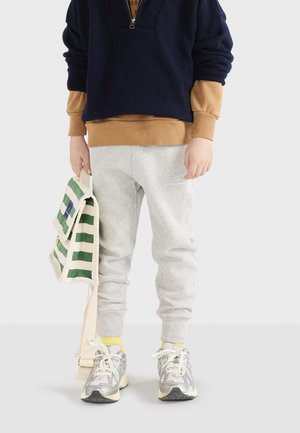 Marinefarbener Pullover, beiger Sweatshirt darunter, graue Sweatpants, gelbe Socken, graue Sneakers und eine gestreifte grünen und weißen Tragetasche.