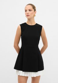 Robe noire sans manches avec un corsage ajusté et une jupe blanche évasée plissée. Tissu lisse, design minimaliste et décolleté rond.