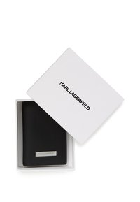 Melns Karl Lagerfeld ādas maks atvērtā baltā kastē ar zīmola nosaukumu uz vāka un metāla logotipa plāksni uz maks priekšpuses.