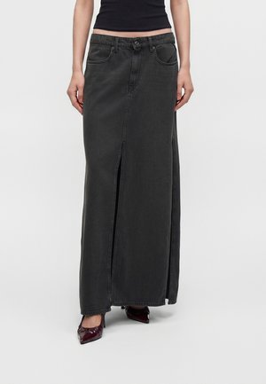 Frau trägt einen hoch taillierten schwarzen Denim-Maxirock mit Frontschlitz, kombiniert mit dunklen spitzen Schuhen und einem kurzen schwarzen Top.
